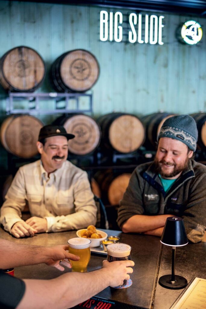 Two men sit at a bar with drinks, smiling as someone hands them beers; barrels are stacked behind them.