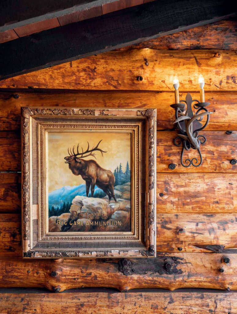Framed elk painting on a log cabin wall next to a rustic metal sconce with two candle-shaped lights.