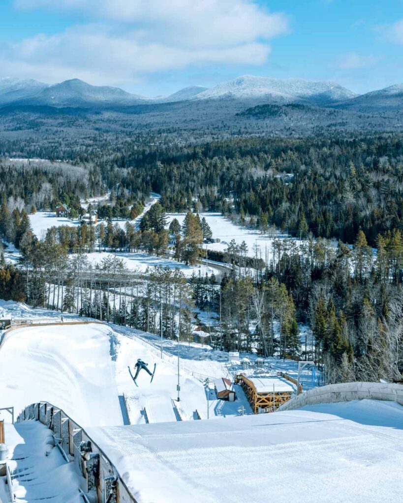 Snowy bobsled track curves through a forested winter landscape with mountains in the background under a blue sky.
