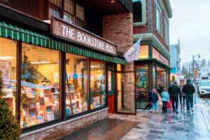 People walk past a brightly lit bookstore on a wet sidewalk in a charming downtown street.