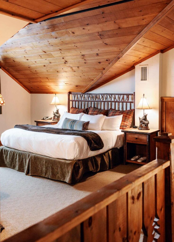 Things to Do in Lake Placid, NY: Where to Eat, Stay, and Play. Cozy attic bedroom with a wood-paneled ceiling, large bed, plaid pillows, and rustic bedside lamps.