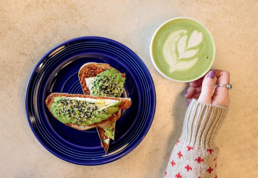 Avocado toast on a blue plate and a green matcha latte with latte art, next to a hand with a ring.