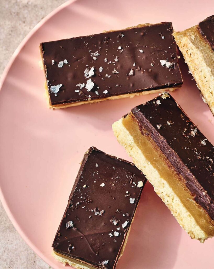 Three chocolate and caramel shortbread bars with sea salt flakes on a pink plate.
