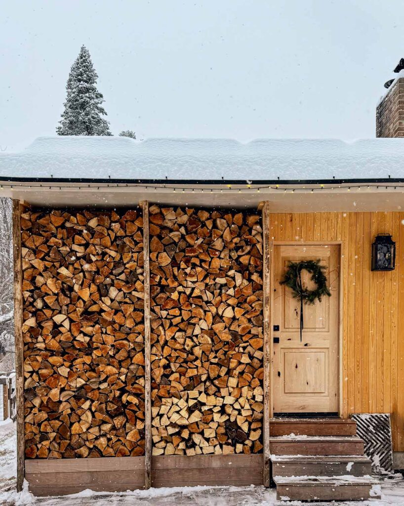 A large stack of firewood next to a wooden door with a wreath, outside a yellow house in snowy weather.