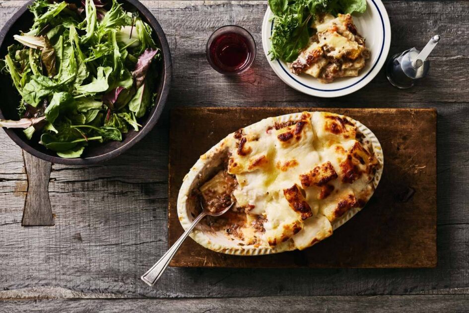 Baked lasagna in a dish with a serving spoon, salad in a bowl, and a plate of salad and lasagna.