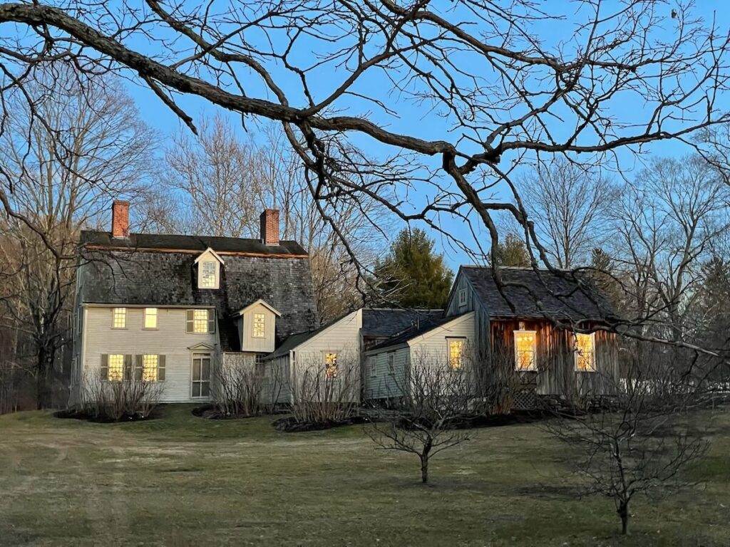 Two connected white houses with lit windows, bare trees, and a twilight sky in early spring.