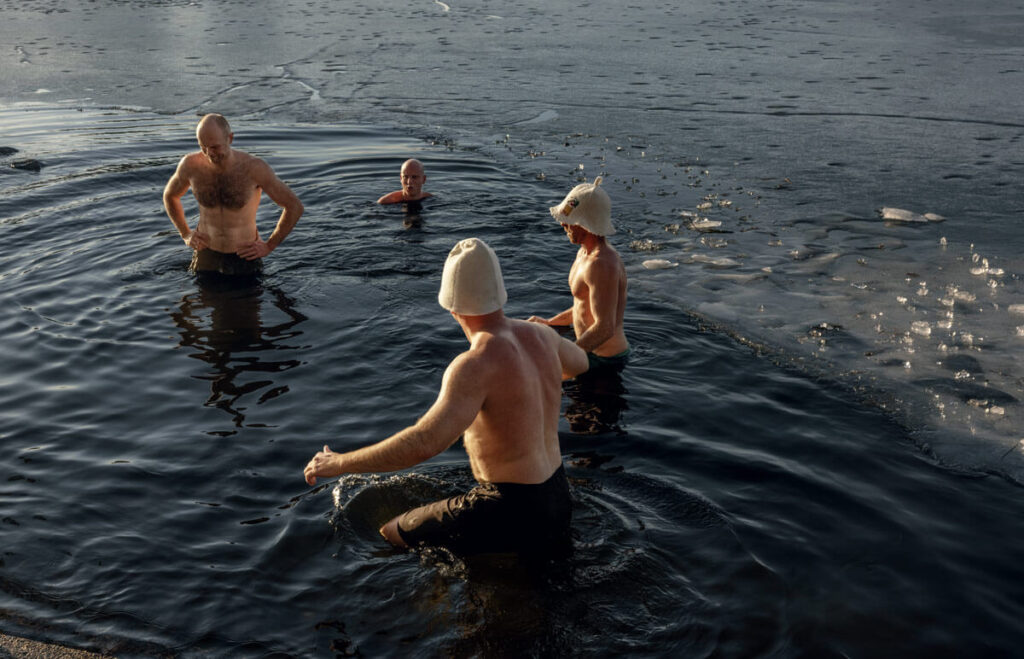 Four men wearing white hats swim in icy water near the edge of a frozen lake on a cold day.