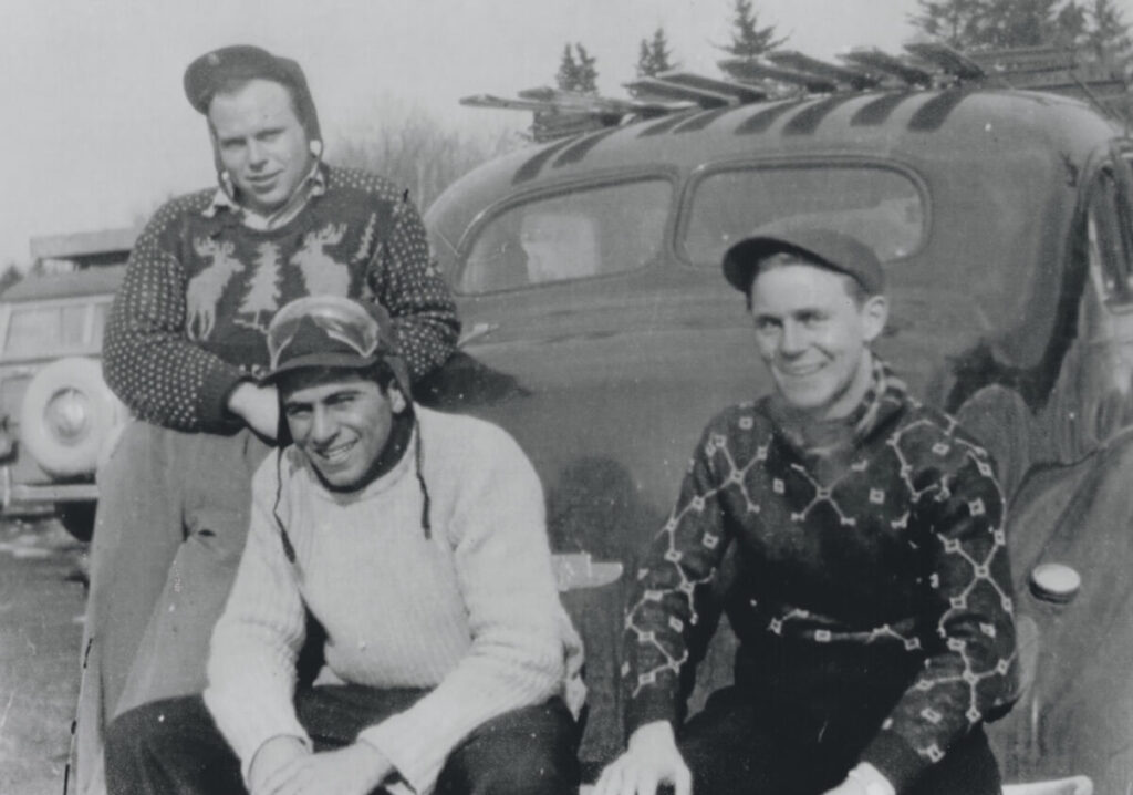 Three men in vintage sweaters and hats pose in front of an old car with skis on the roof, black and white photo.