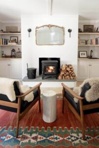 Cozy living room with wood stove, stacked firewood, two chairs, and a decorative mirror above the fireplace.