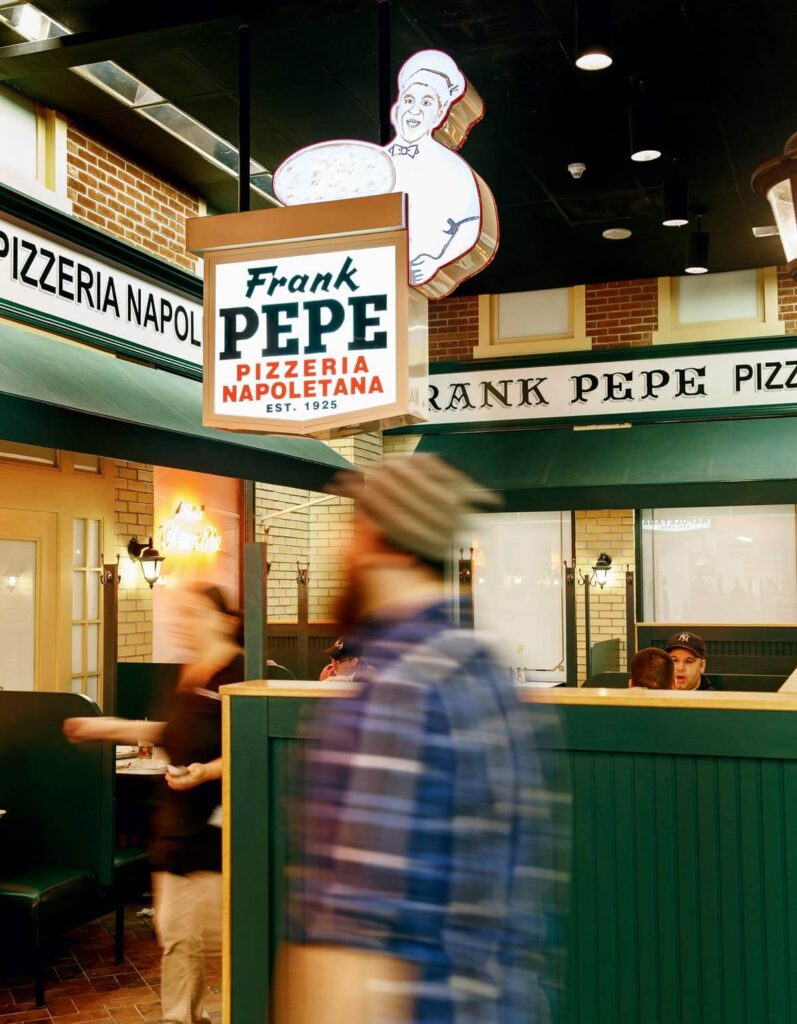 People walking inside Frank Pepe Pizzeria Napoletana, with a large pizza chef sign above the entrance.