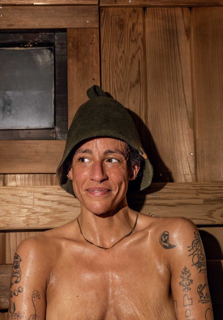 A smiling person with tattoos wears a dark sauna hat and sits in a wooden sauna, skin glistening with sweat.