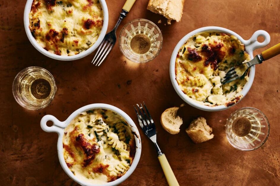 Three bowls of baked pasta with melted cheese, bread pieces, and glasses of white wine on a brown table.