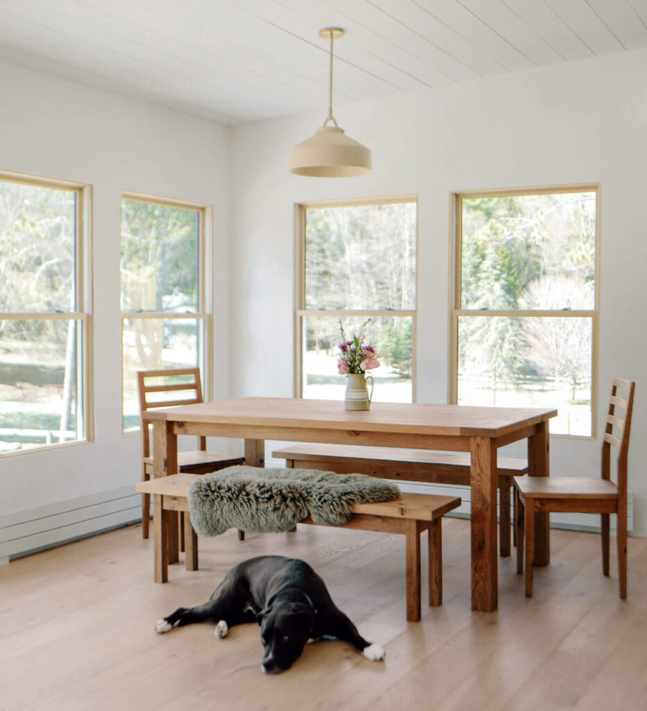Bright dining room with wooden table, chairs, bench, vase of flowers, and a black dog lying on the floor.