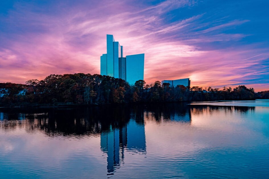 Tall modern buildings reflect on a calm lake at sunset, with vibrant pink and blue clouds in the sky.
