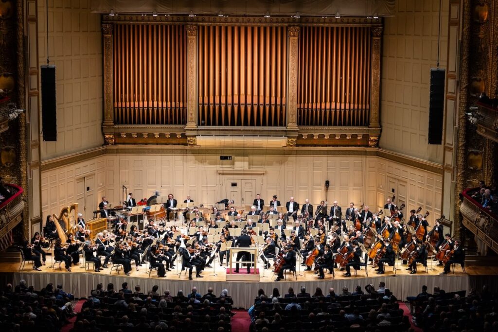 A full symphony orchestra performs on stage in a grand concert hall before an audience. BSO American music festival