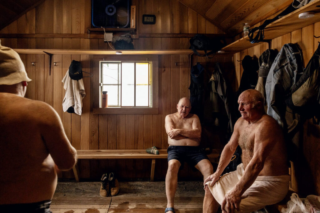 Three older men sit in a wooden sauna, two wrapped in towels, with clothes and boots hanging on the walls.