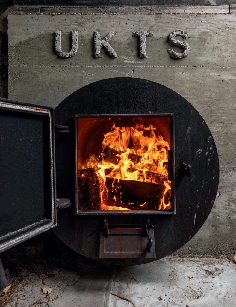 A round metal furnace with an open door, burning logs inside; “UKTS” is on the wall above.