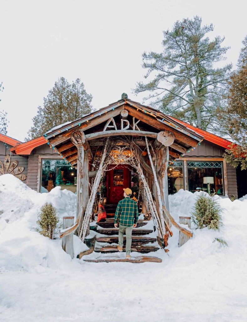 Person in winter clothes walks up snowy steps to a rustic wooden cabin surrounded by trees.