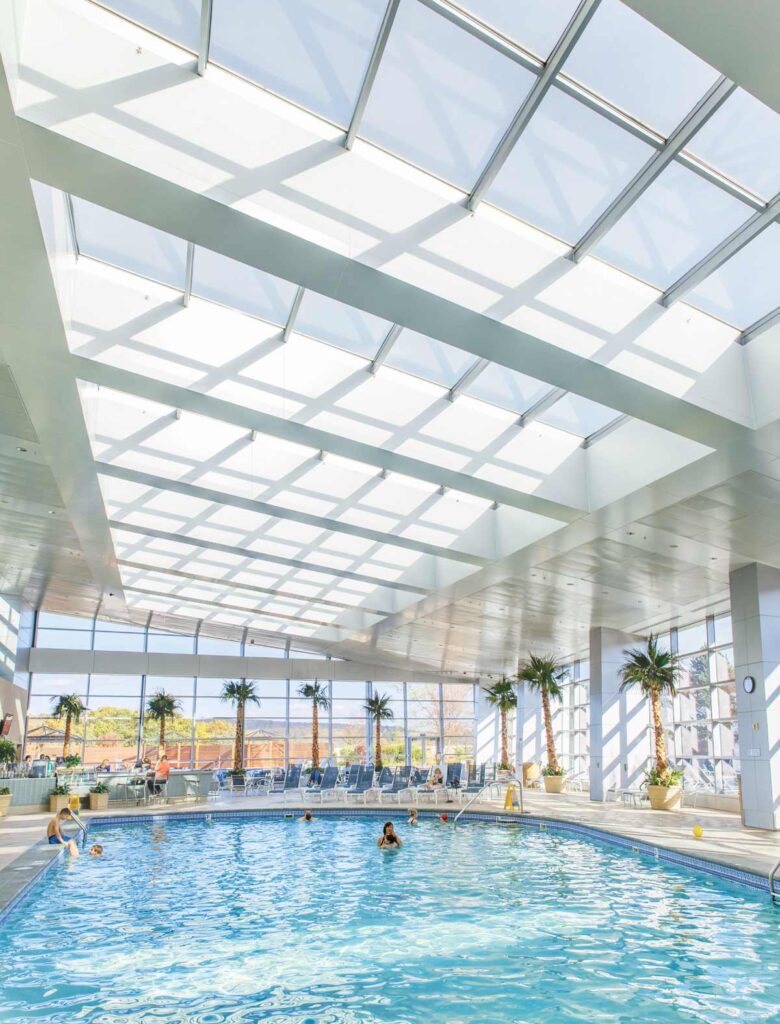 Indoor pool with people swimming under a large glass ceiling and surrounded by tall windows and palm trees.