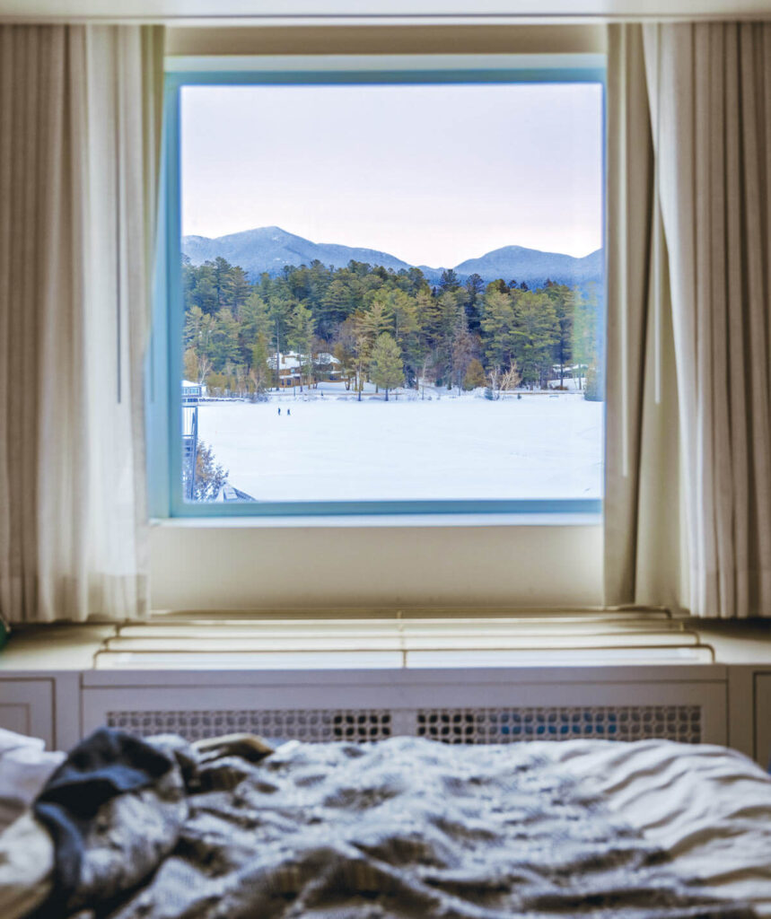 Snowy landscape with trees and mountains, viewed through a window from a cozy bed with rumpled blankets.