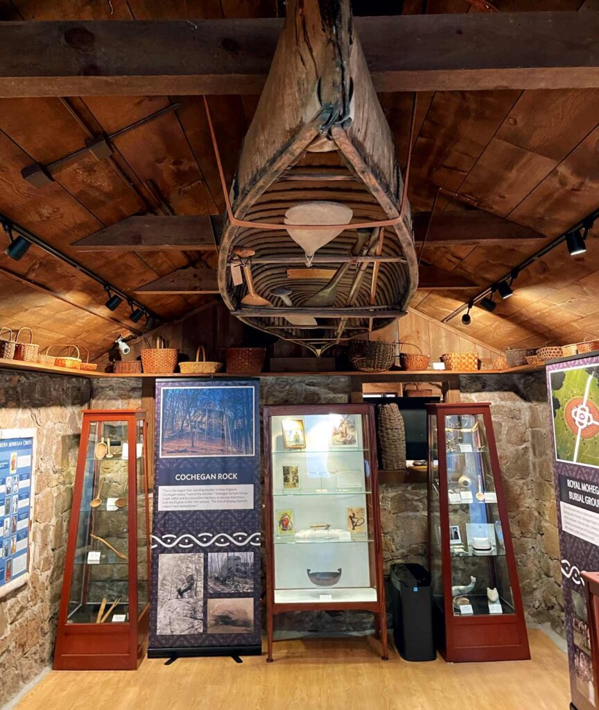 A wooden canoe hangs from a rustic museum ceiling above display cases with artifacts and informational panels.