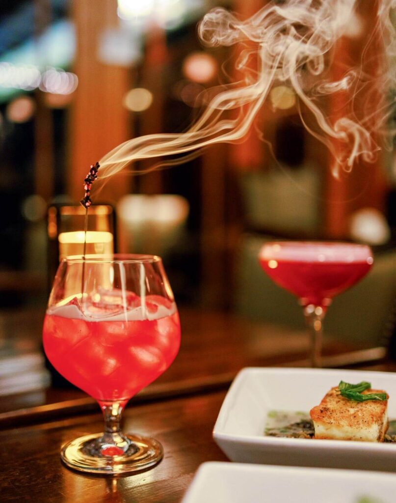 A pink cocktail with ice in a glass, garnished with smoldering herbs releasing smoke, sits on a table.
