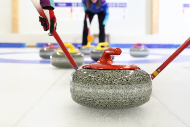 Close-up of curling stones on ice with players sweeping in the background during a curling match.