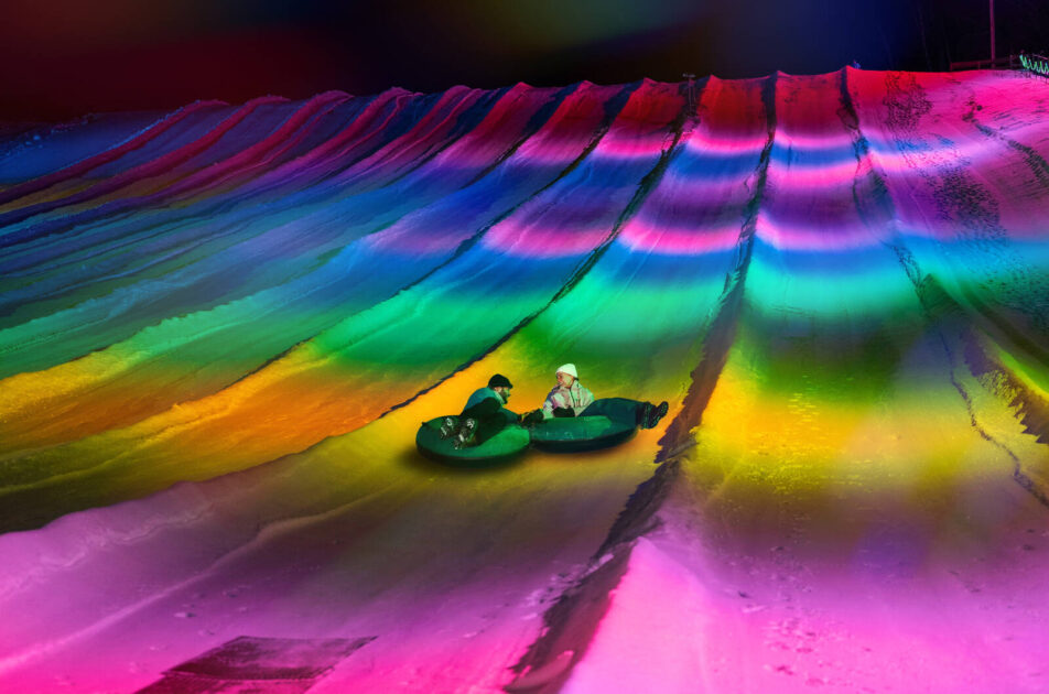 Arctic Tubing at Cranmore Mountain Resort in New Hampshire - Two people tubing down a snow hill illuminated with bright, colorful rainbow lights at night.