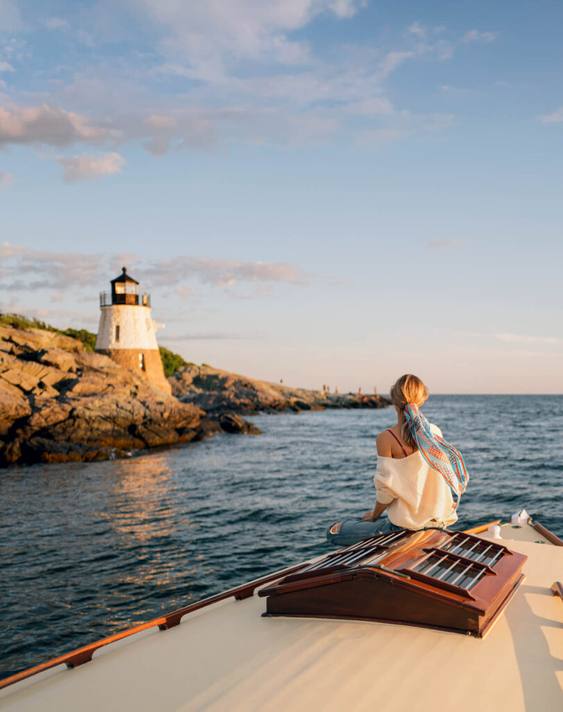 A woman sits on a boat at sunset, facing a rocky lighthouse by the sea.