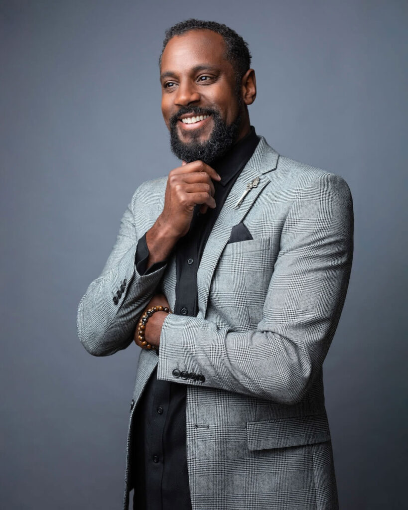 Smiling man in a gray suit with a black shirt, posing with hand on chin against a gray background.