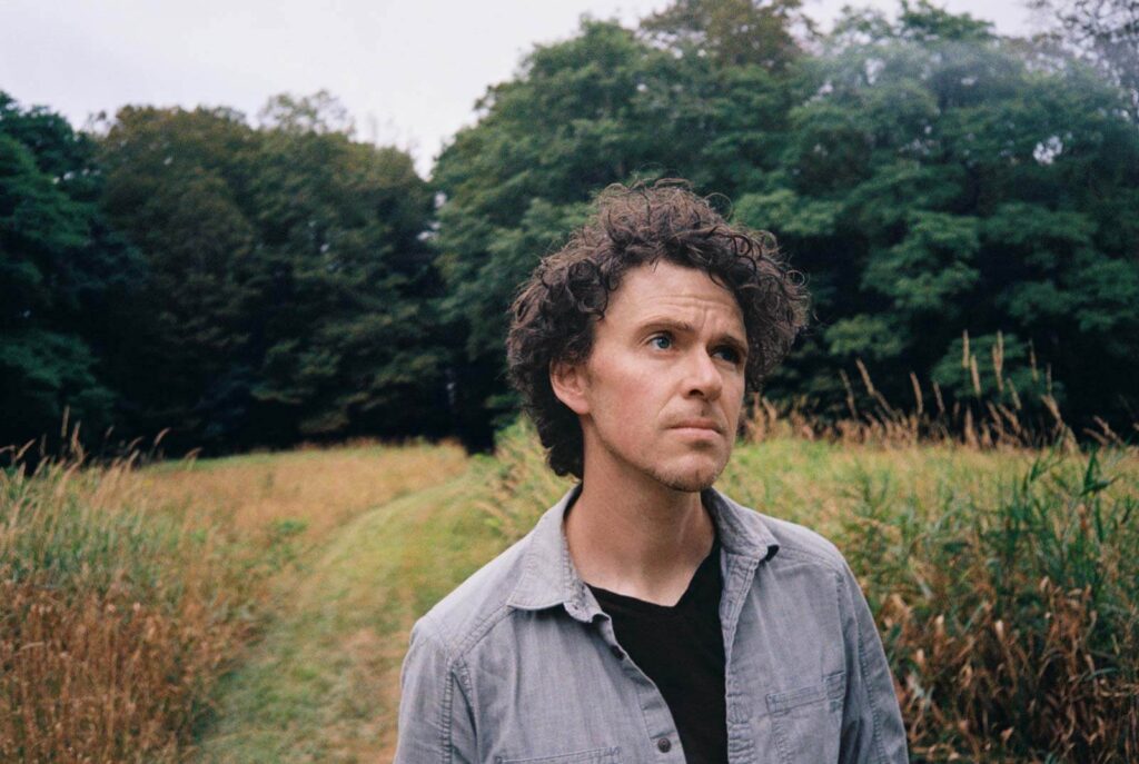 A man with curly hair stands in a grassy field, looking thoughtfully upward, with trees in the background.