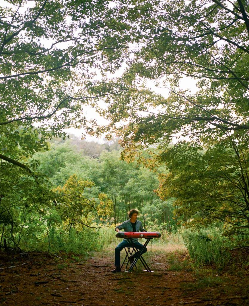 Ben Cogrove plays a keyboard on a stand in a lush green forest clearing under leafy tree branches.