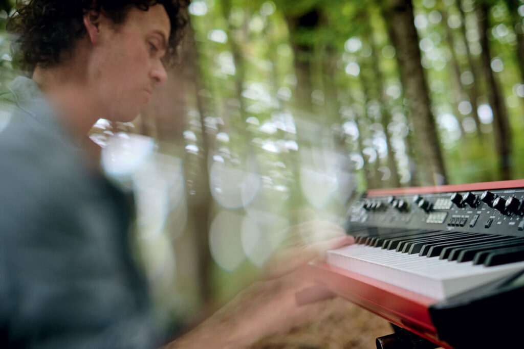 A person plays a keyboard outdoors, surrounded by blurred trees and soft natural light.