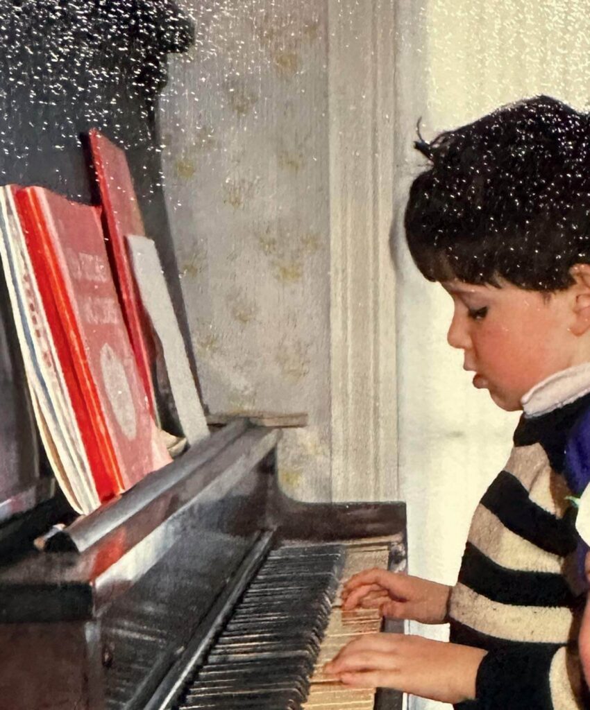 Young child playing piano with sheet music on the stand, wearing a striped sweater, viewed from the side.