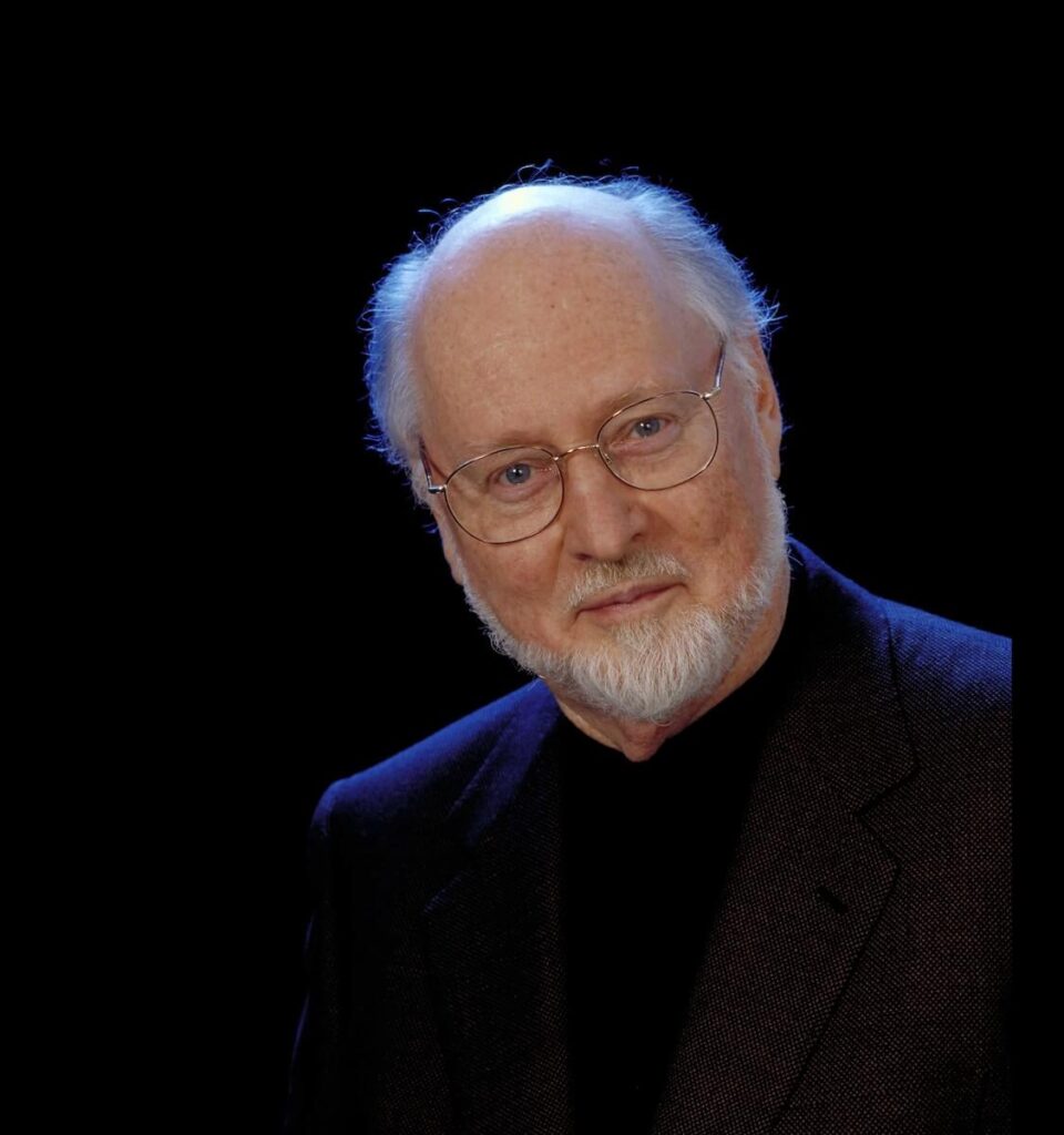 Conductor John Williams, an elderly man with glasses and a white beard, wearing a dark suit, poses against a black background.