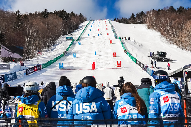 Spectators in winter gear watch a ski competition on a snowy slope with moguls and gate markers.