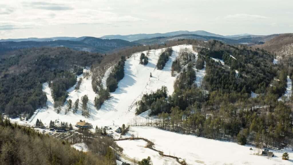 Aerial view of a snowy ski slope on a forested mountain with surrounding hills and a few buildings below.