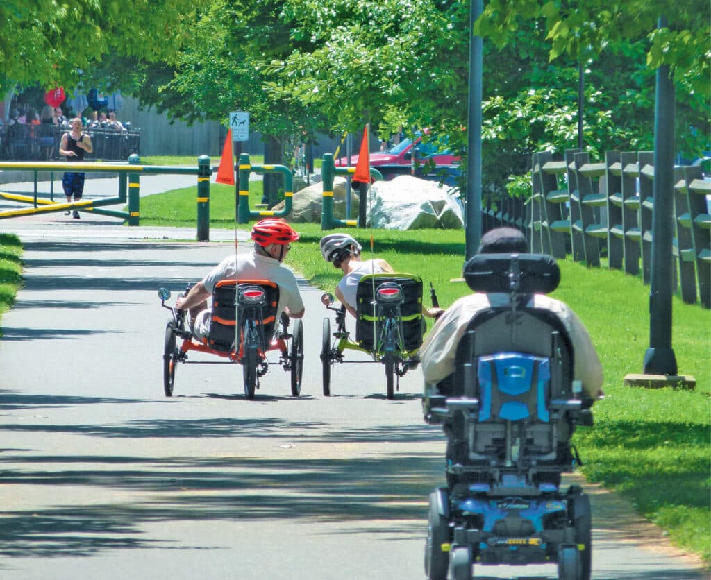 Three people in adaptive bikes and a wheelchair travel down a sunny, tree-lined park path.