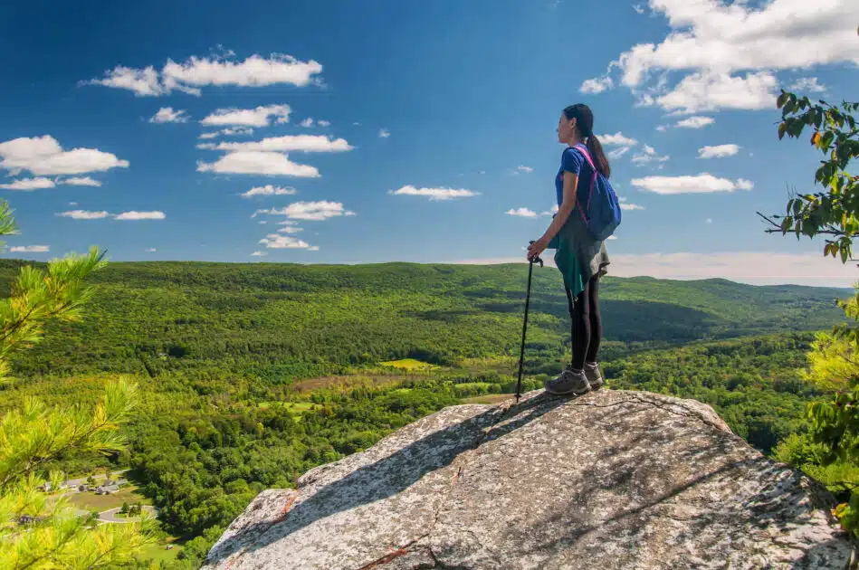 A hiker with a backpack stands on a rocky cliff overlooking a lush green valley under a blue sky.