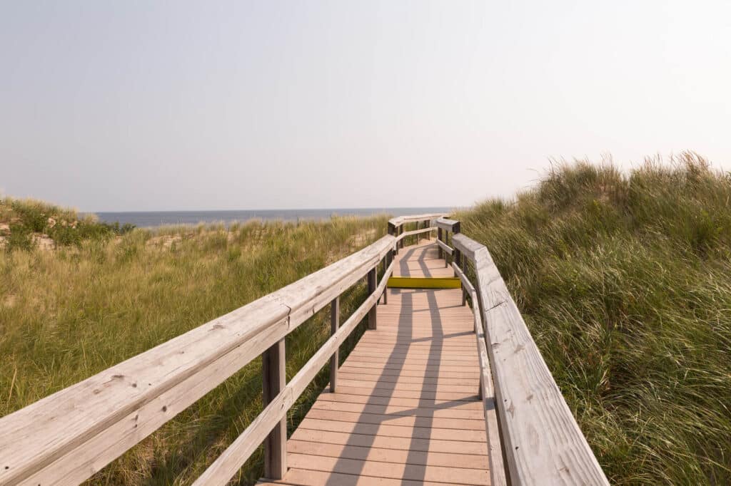 Wooden boardwalk winding through grassy dunes toward the beach under a clear sky.