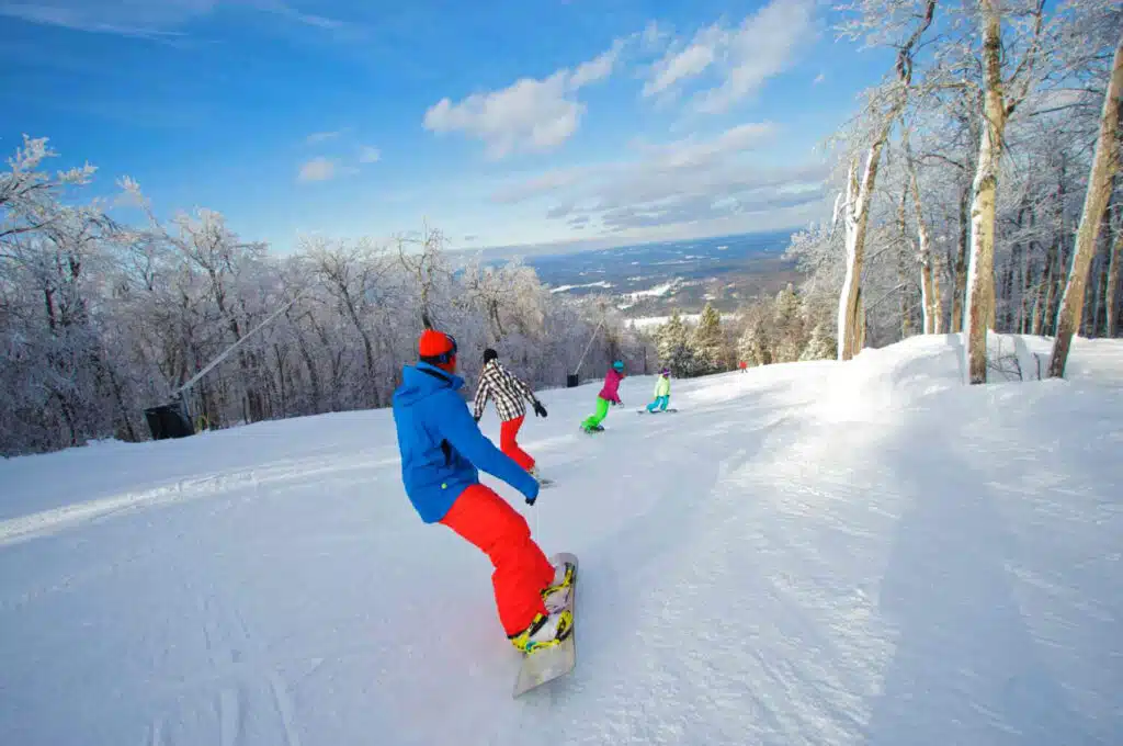 Four people snowboarding down a snowy, tree-lined slope under a bright blue sky.
