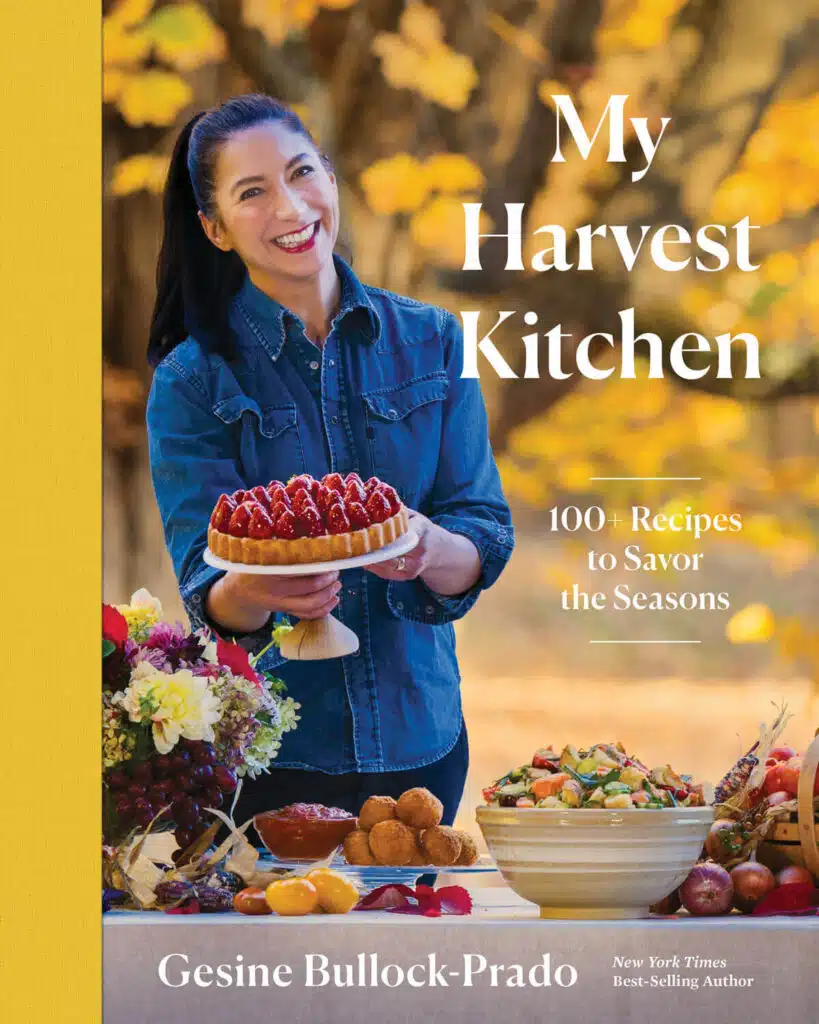 A smiling woman holds a berry tart outside by a table with flowers, fruit, and autumn leaves in the background; cover for "My Harvest Kitchen" cookbook.