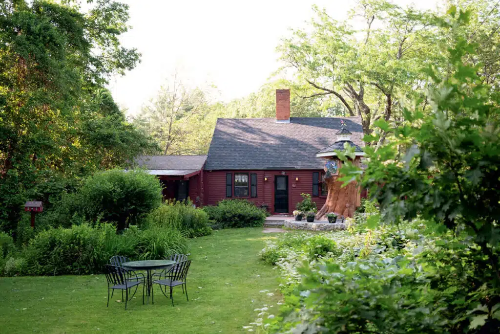 A cozy red cottage with a lush green garden, patio chairs, and trees surrounding the house.