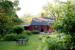 A cozy red cottage with a lush green garden, patio chairs, and trees surrounding the house.