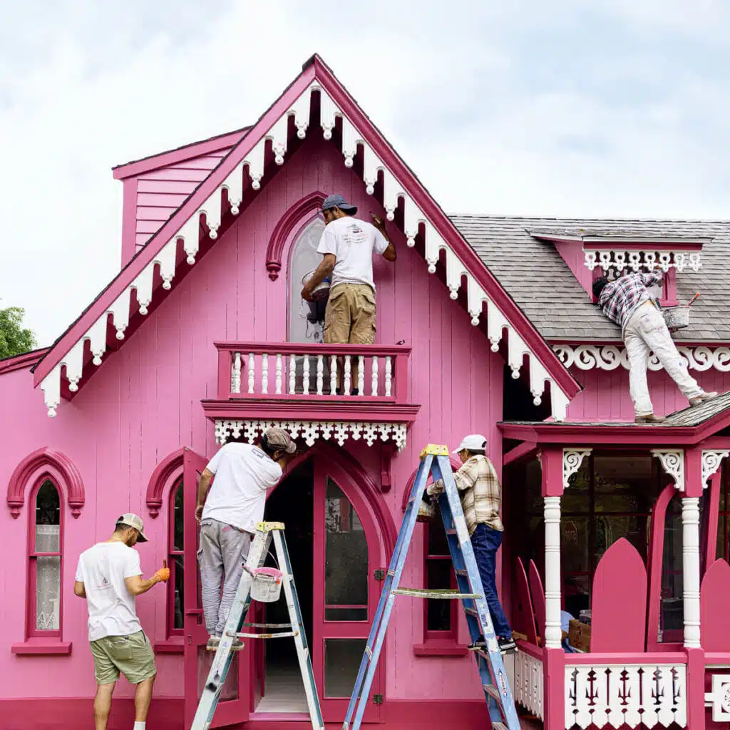 New England Victorian Gingerbread Houses. Four people paint a detailed, bright pink Victorian house using ladders and brushes on a sunny day.