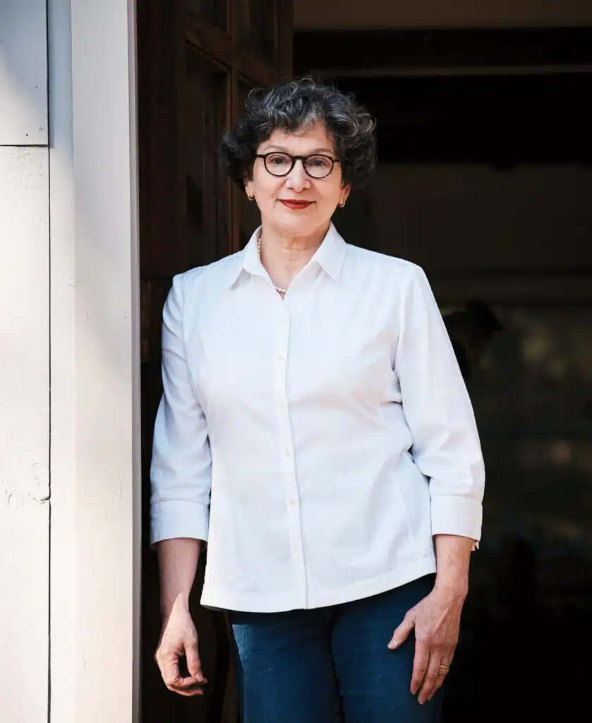 Connecticut ceramic artist Frances Palmer. Woman with short curly hair, glasses, and a white shirt stands smiling in a doorway, hand at her side.
