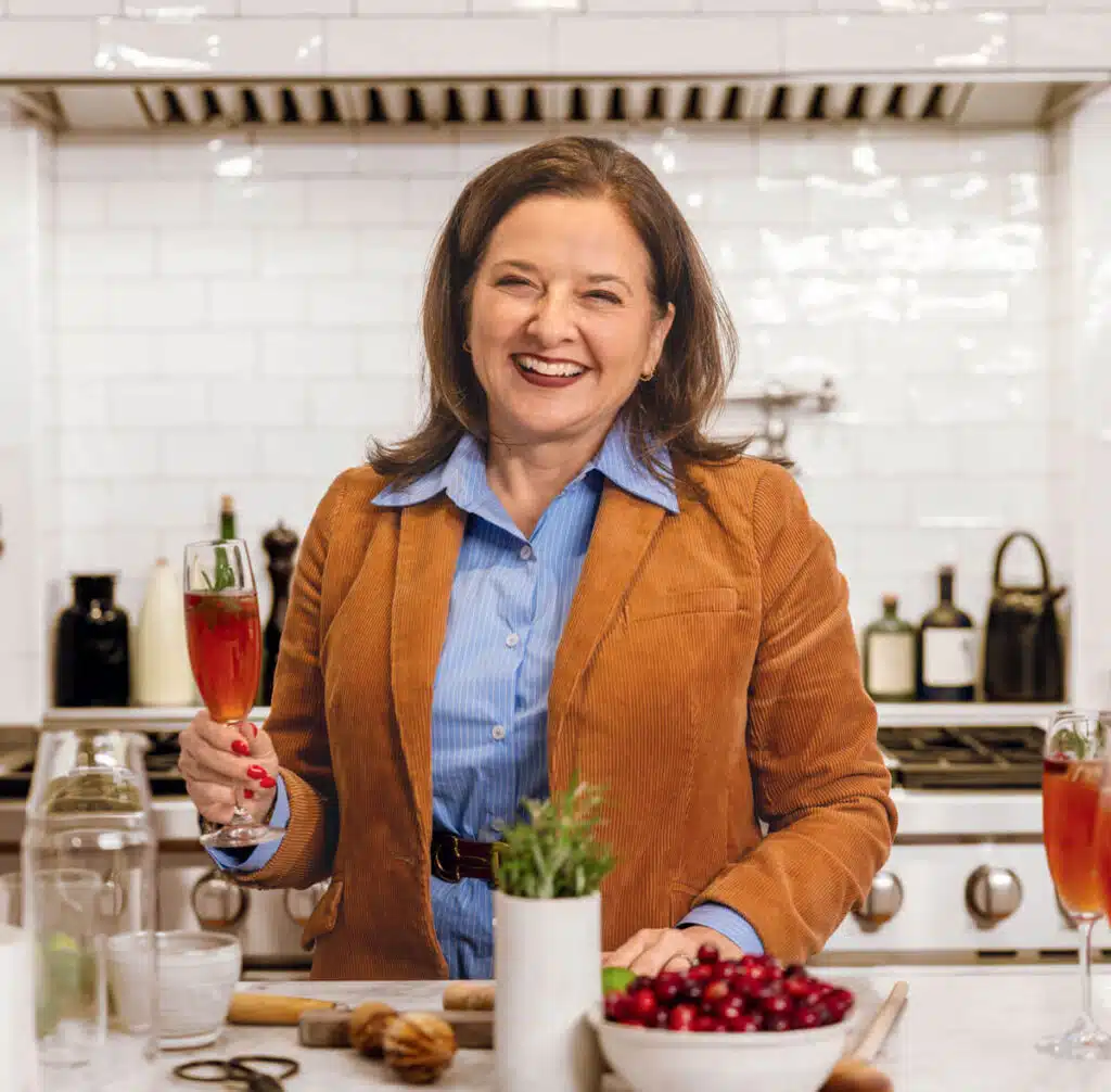 Smiling woman in a kitchen holding a glass of red drink, with ingredients and utensils on the counter.