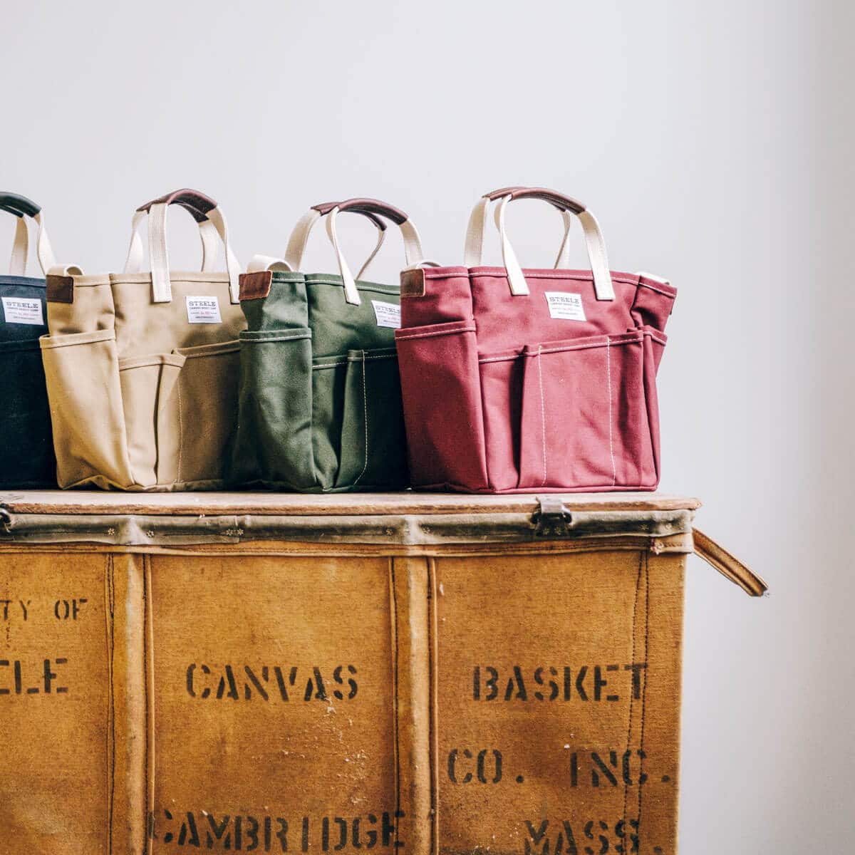 Four canvas tote bags in black, beige, green, and red sit on a vintage wooden chest against a white wall.