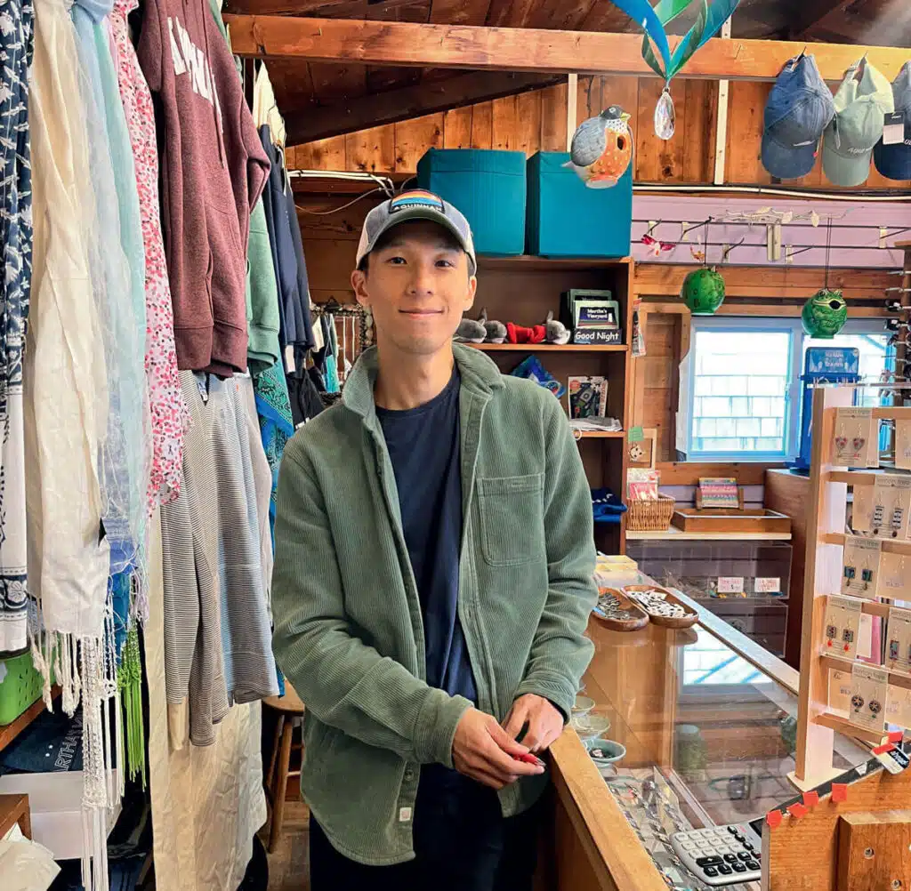 Conversations | Wampanoag Writer Joseph Lee. A person in a green jacket and cap stands smiling inside a colorful shop with clothes and accessories.