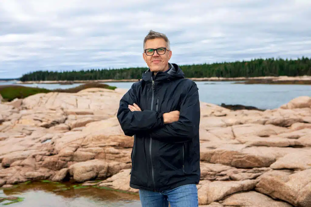 A man in a black jacket stands on rocky terrain by water, with trees and cloudy sky in the background.